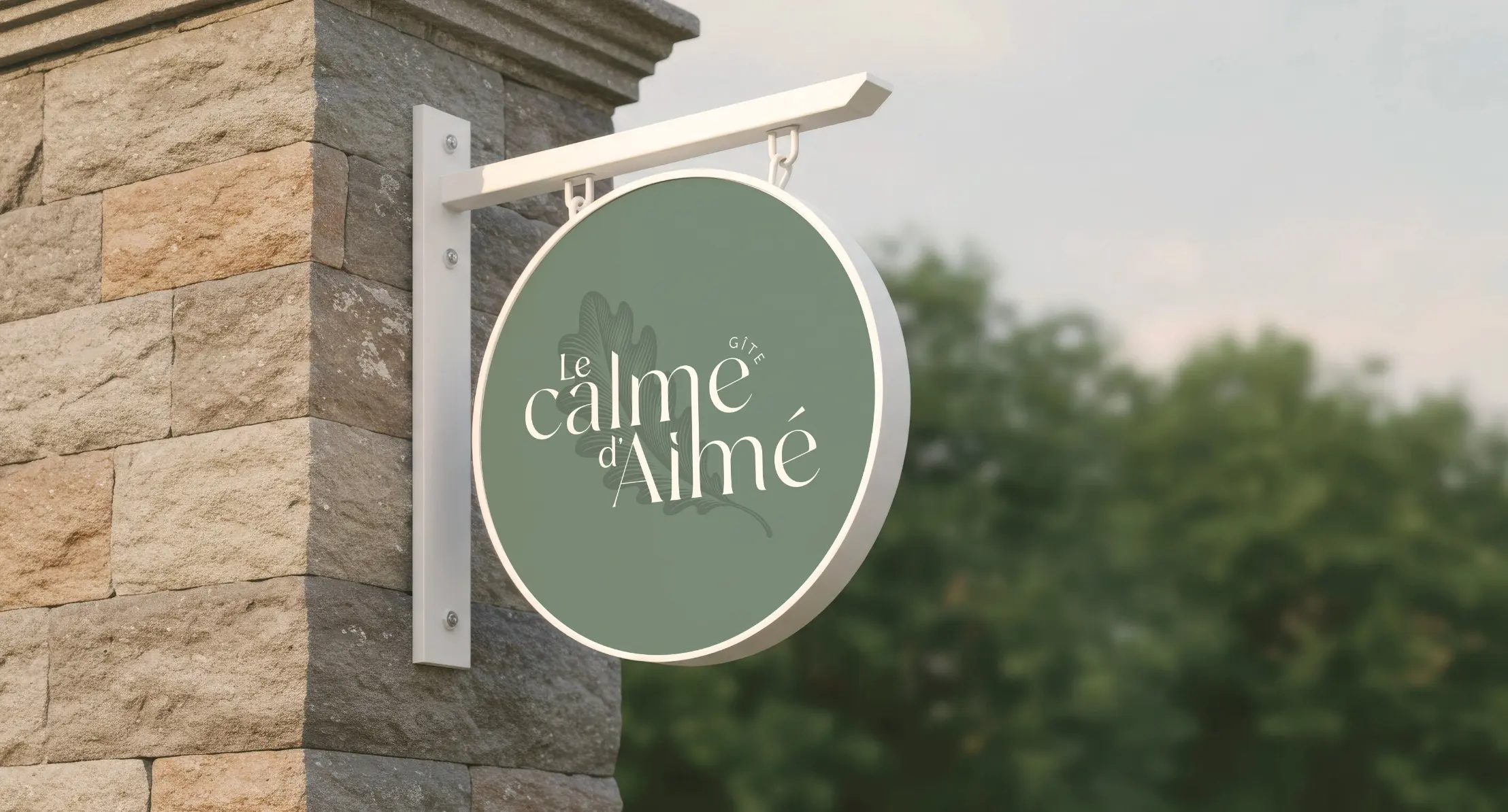Mockup d'enseigne ronde murale Le Calme d'Aimé Gîte en Vendée, fond vert sauge avec logo blanc sur mur en pierre, identité visuelle conçue par Tchak Studio